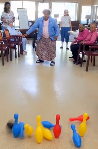 An elderly woman is bowling with colorful plastic pins set up on the floor in a well-lit room. Several other elderly individuals and two caregivers are seated or standing around, watching and smiling. The room has large windows and chairs arranged along the walls.