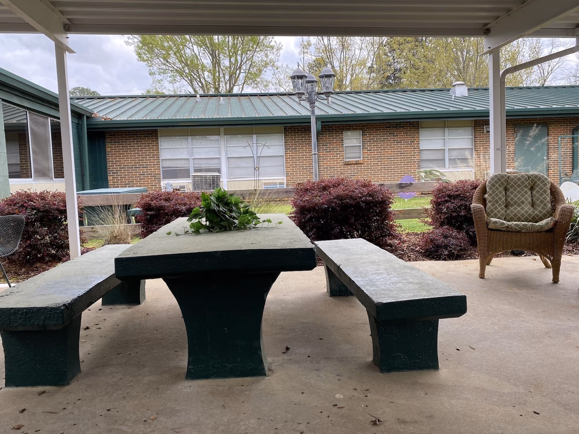 Covered outdoor seating area with a concrete table and two concrete benches, a wicker chair with a cushion, and some greenery on the table. In the background, there is a brick building with green metal roofing and windows, surrounded by bushes and trees.