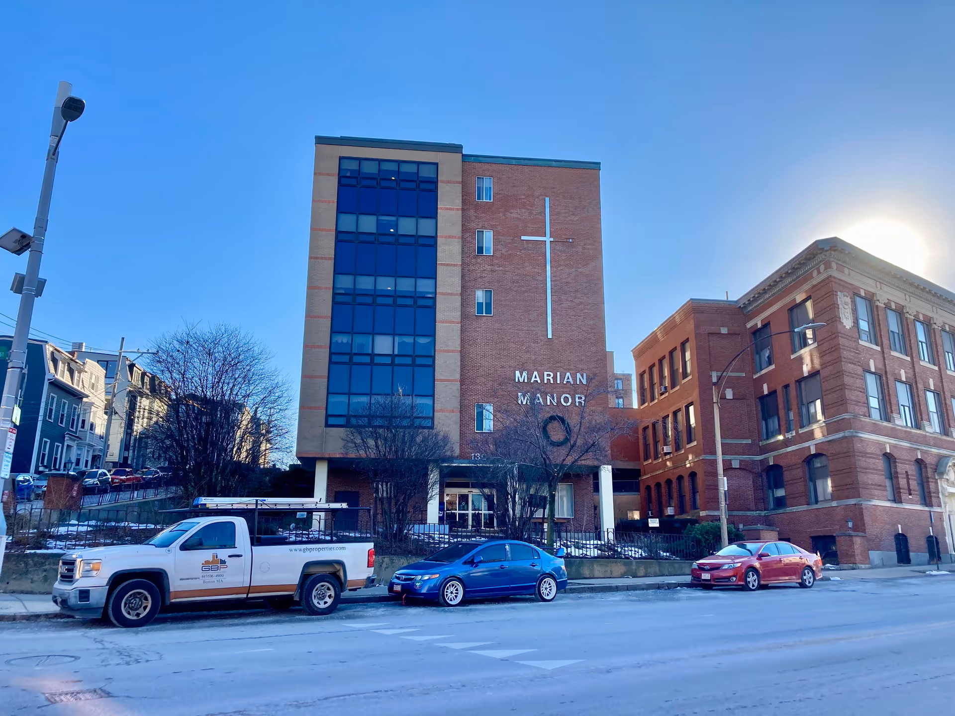 Exterior view of Marian Manor Nursing Home, a multi-story brick building with large windows and a prominent cross on the facade. Several cars are parked along the street in front of the building under a clear blue sky.