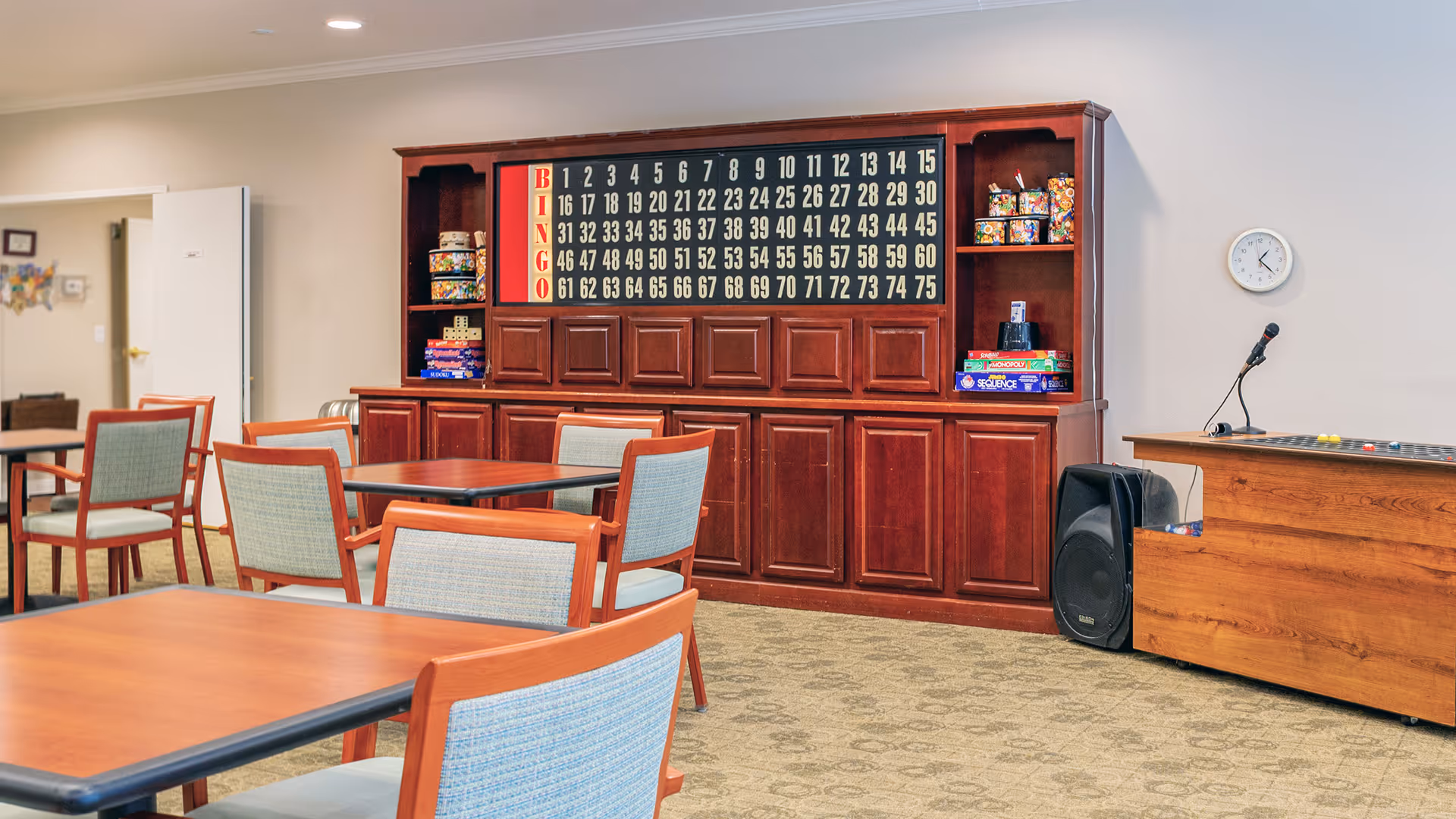 A community room with several wooden tables and chairs arranged for group activities. A large wooden cabinet against the wall holds a bingo board with numbers and the word BINGO displayed vertically. Shelves on the cabinet contain board games and bingo supplies. To the right, there is a wooden podium with a microphone and a speaker on the floor. A clock is mounted on the wall above the podium.