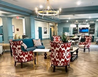 Spacious senior living common room with a teal sofa and red patterned armchairs around coffee tables, chandeliers overhead, and a dining area with a TV and built-in cabinets in the background.