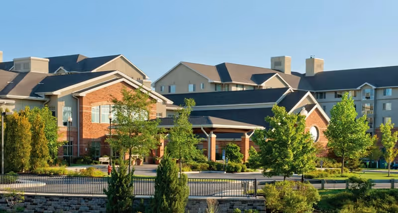 Exterior view of Brooksby Village Senior Living Community showing multiple connected buildings with brick and beige siding, surrounded by green trees and landscaping under a clear blue sky.