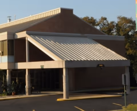 Exterior view of a building entrance with a large white awning supported by columns, surrounded by trees and a parking area.