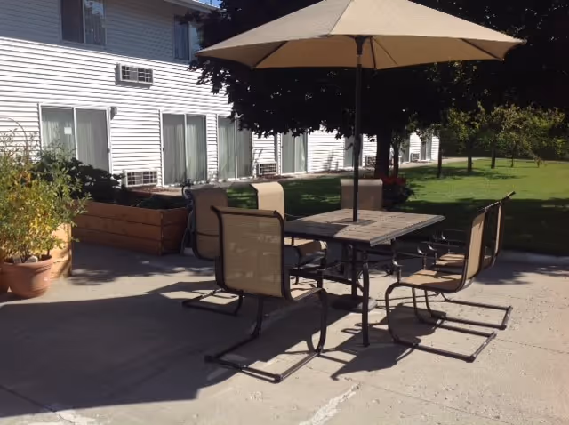 Outdoor patio area with a table, four chairs, and a large beige umbrella providing shade. The patio is adjacent to a white building with sliding glass doors and windows. There are potted plants and a raised garden bed near the building, and a grassy area with trees in the background.