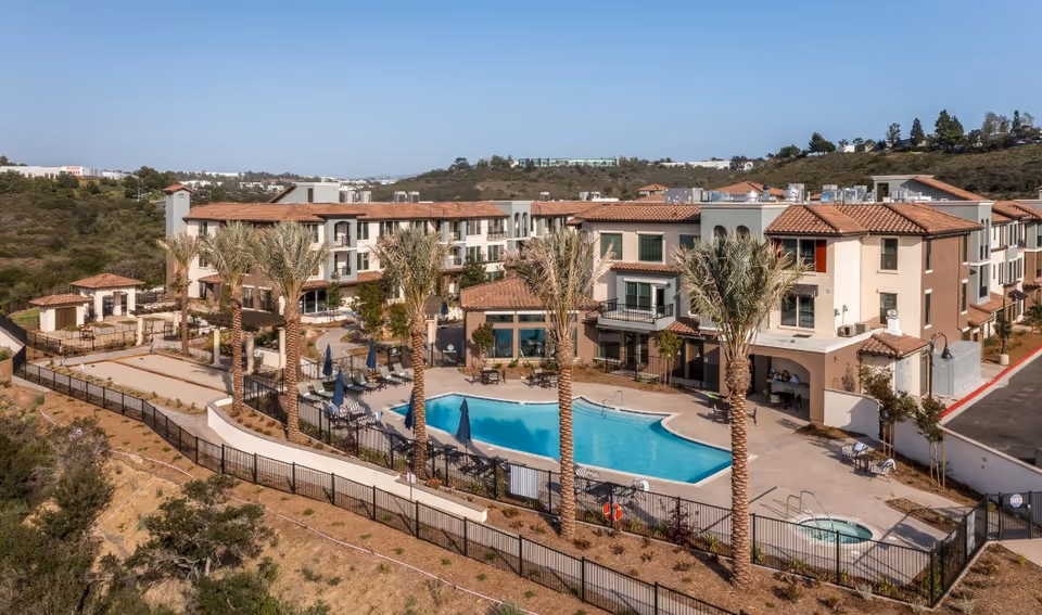 A multi-story senior living complex with a large outdoor pool, palm trees, and patio seating.