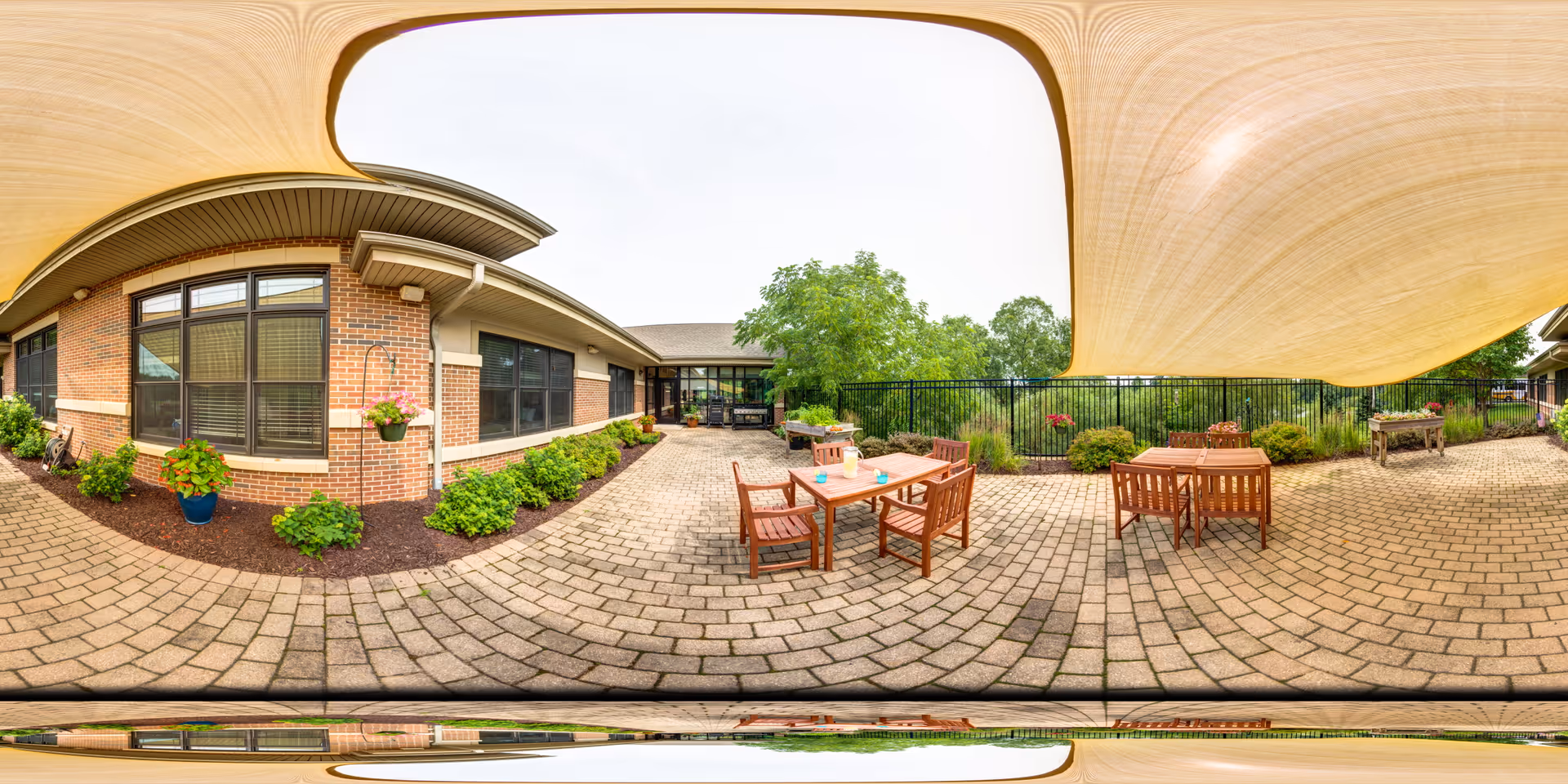 Shaded brick-paved courtyard with wooden tables and chairs, planters, and a low brick building facade under a large fabric canopy.