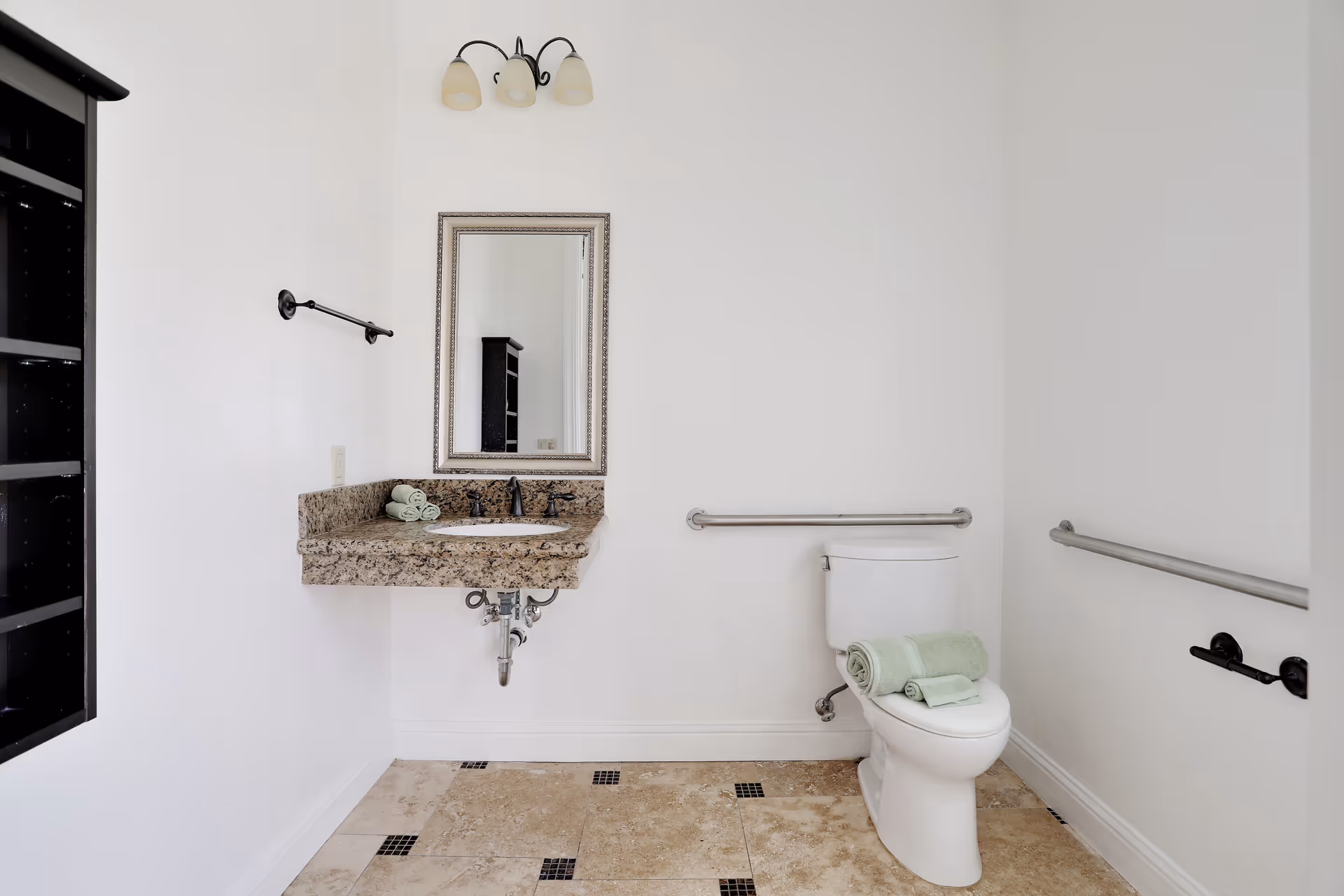 A clean bathroom with a white toilet and a granite countertop sink. The toilet has neatly folded green towels placed on top. There are stainless steel grab bars on the walls beside and behind the toilet. A rectangular mirror with a decorative frame hangs above the sink, and a three-light fixture is mounted above the mirror. The floor is tiled with beige tiles accented by small black square tiles. A black shelving unit is partially visible on the left side of the image.