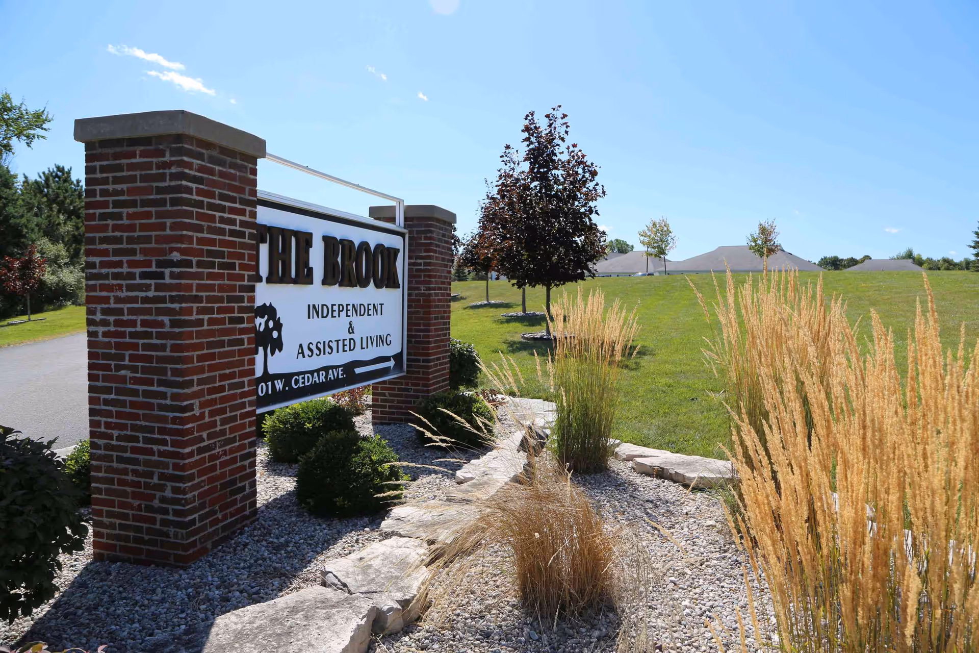 Brick entrance sign for The Brook Independent & Assisted Living amid landscaped rocks, ornamental grasses and a green lawn under a blue sky.