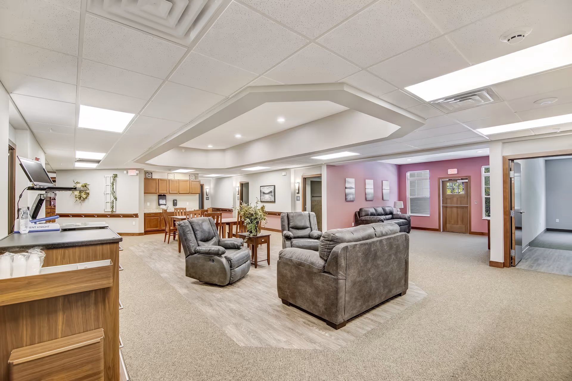 A spacious senior living common area with gray leather sofas and armchairs arranged around a small wooden table with a plant. In the background, there is a dining area with wooden tables and chairs, and a kitchenette with cabinets. The walls are painted in neutral tones with one accent wall in purple. The ceiling has recessed lighting and a drop ceiling design. There is a wooden door and windows letting in natural light.