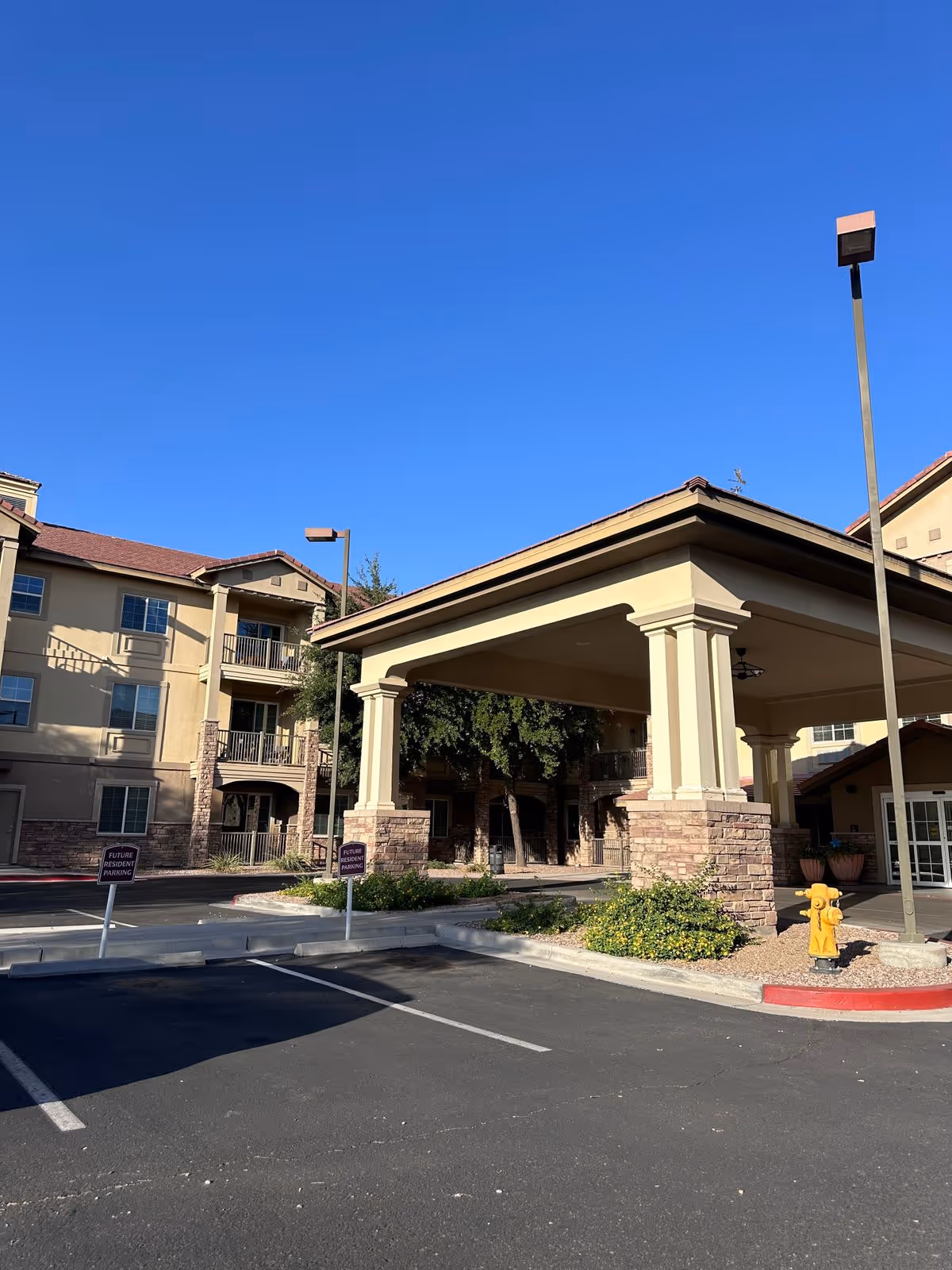 Drive-up entrance and porte-cochere of a senior living building with parking spaces, lamp posts, and a clear blue sky.