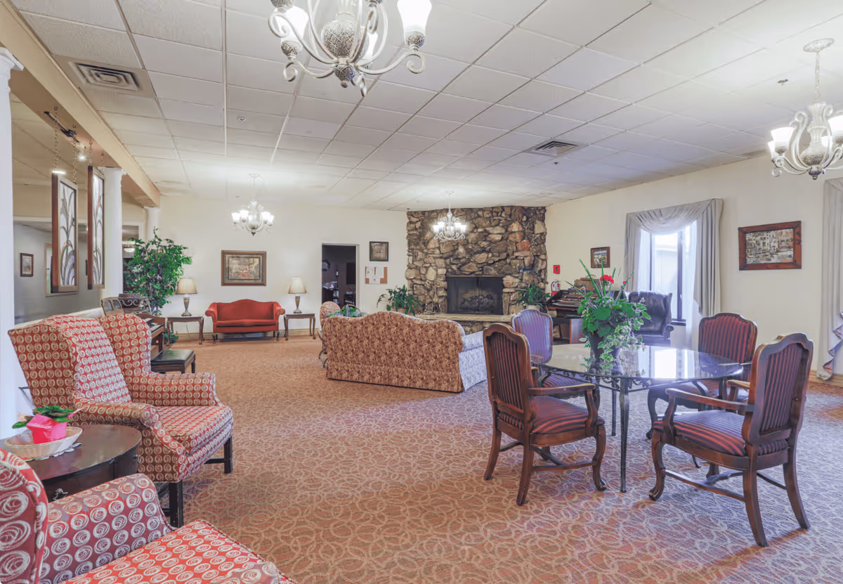 A spacious senior living facility common area with patterned carpet and multiple seating arrangements including upholstered armchairs, sofas, and wooden chairs around a glass table. The room features a stone fireplace, chandeliers, framed artwork on the walls, potted plants, and large windows with drapes allowing natural light.
