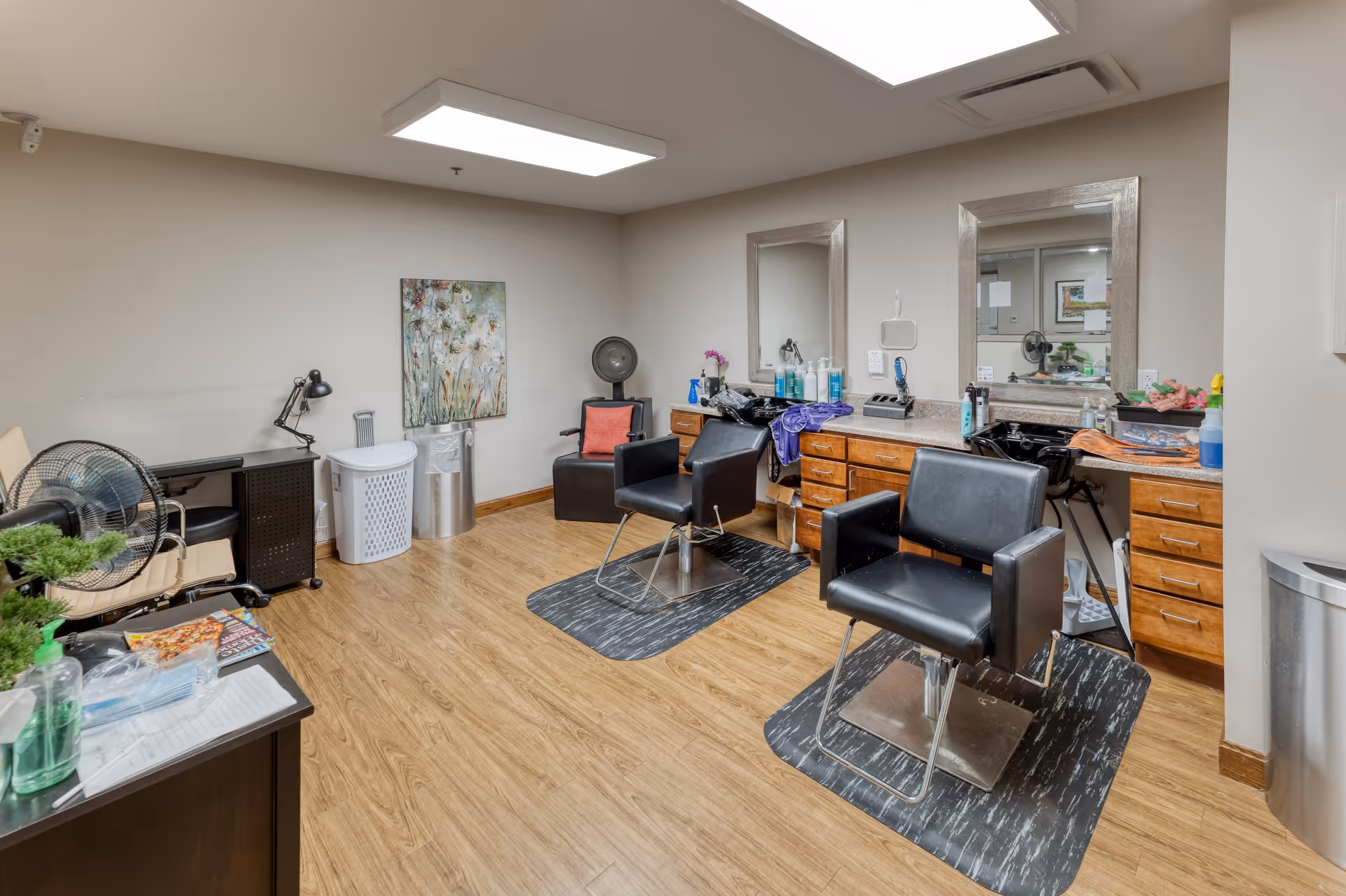 Interior view of a salon area in a senior living facility with two black salon chairs in front of a counter with two mirrors. The counter has various hair care products and towels. The room has wood flooring, a painting on the wall, and additional seating and fans in the corner.