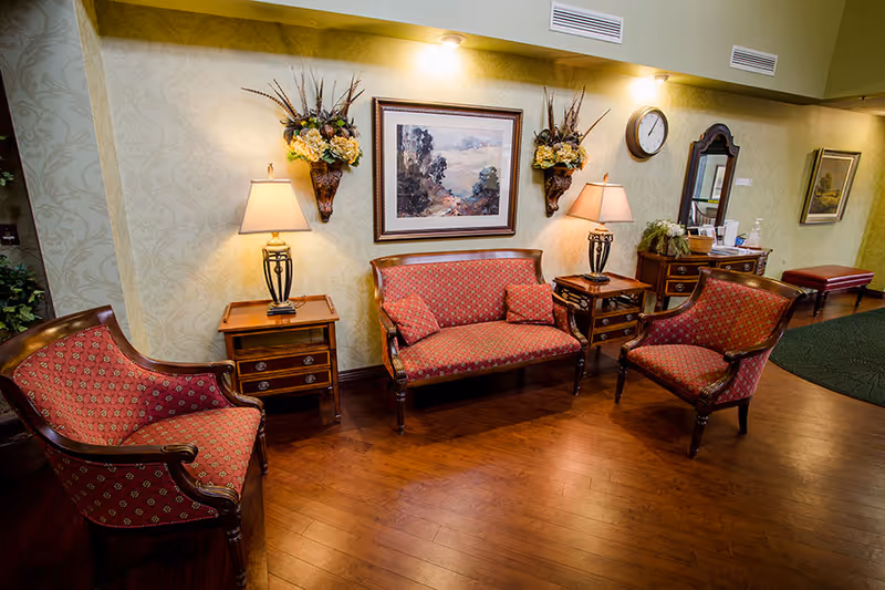 A cozy sitting area with vintage-style furniture including a red patterned loveseat and two matching armchairs. There are two wooden side tables with lamps on either side of the loveseat, floral wall sconces, a framed landscape painting, a wall clock, and a wooden dresser with a mirror. The floor is wooden and the walls have a light patterned wallpaper.