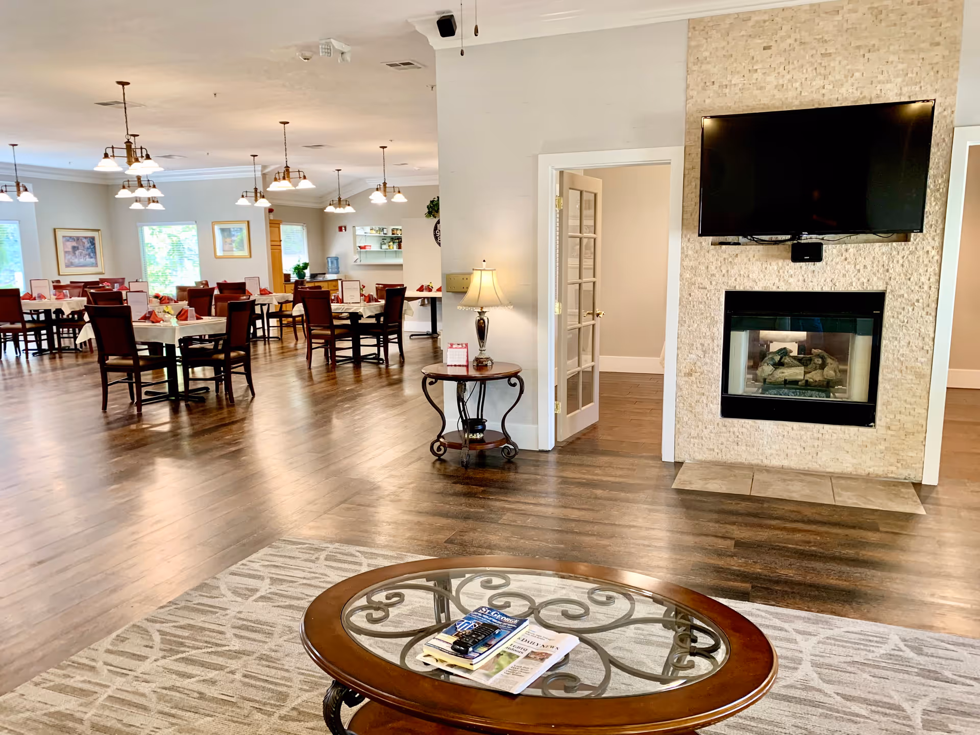 Interior view of a senior living facility showing a spacious dining area with multiple tables and chairs set with napkins and menus. In the foreground, there is a glass-top coffee table with magazines and a remote control on a patterned rug. To the right, a wall features a mounted flat-screen TV above a modern fireplace. A small side table with a lamp is positioned near a doorway with glass-paneled double doors.