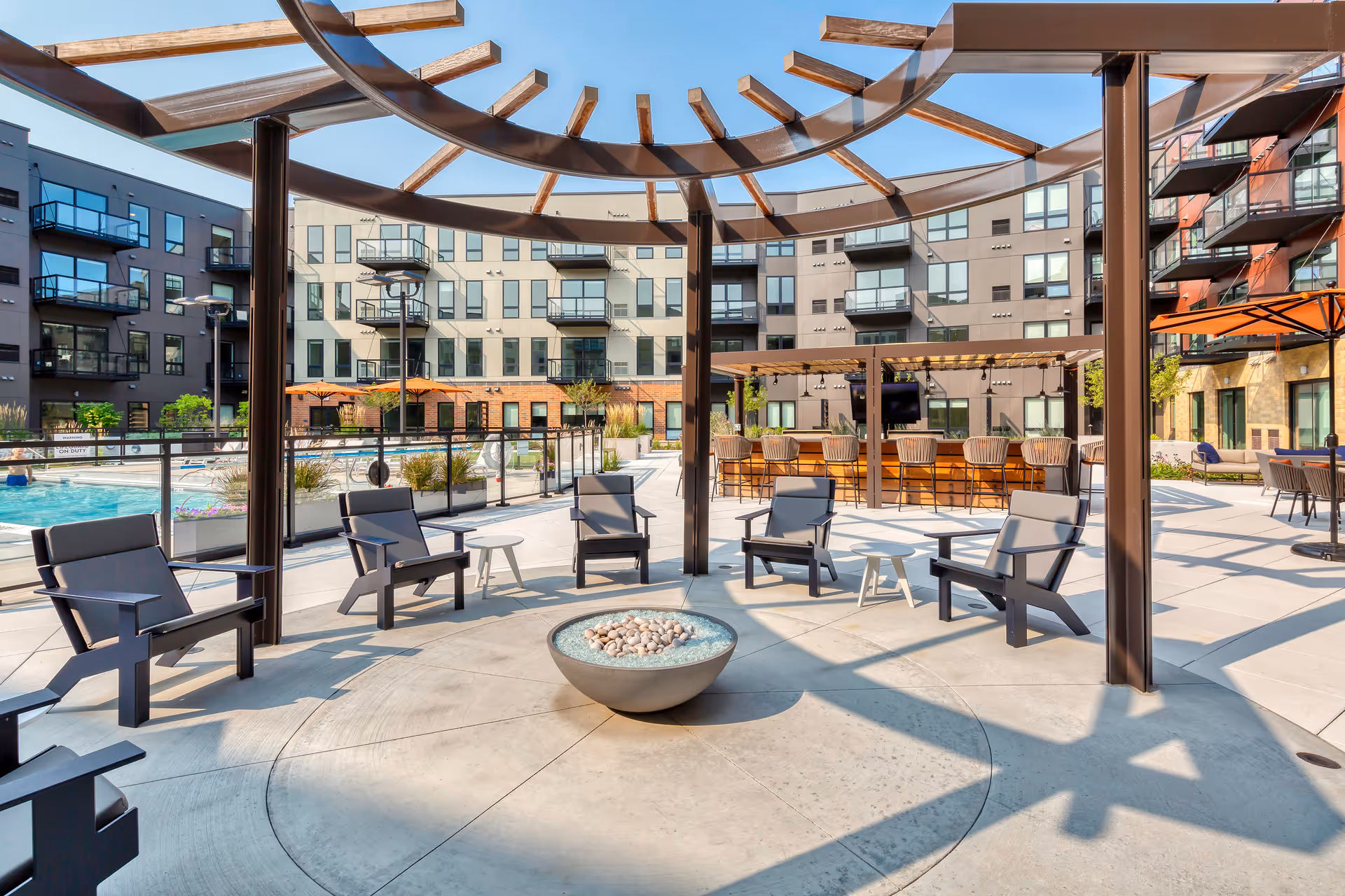 Outdoor seating area at Avidor Glenview featuring a circular arrangement of black chairs around a modern fire pit under a pergola with wooden beams. In the background, there is a swimming pool with lounge chairs and umbrellas, a bar area with stools, and a multi-story apartment building with balconies.