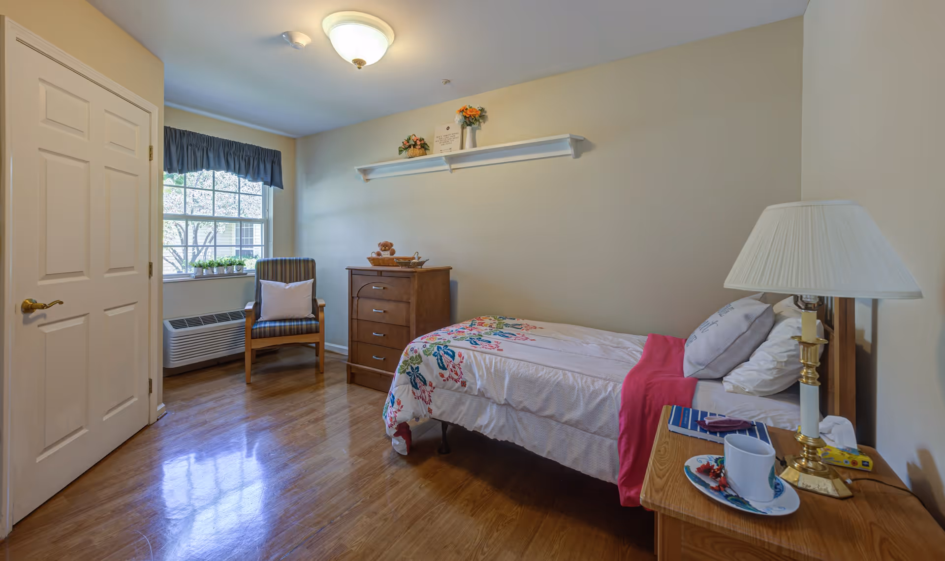 A small, neatly arranged bedroom with a single bed covered in a floral bedspread and a pink blanket. There is a wooden nightstand with a lamp, a cup on a saucer, and some personal items. A wooden dresser with a basket and decorative items sits against the wall. A wooden chair with a cushion is placed near a window with a blue valance, and the floor is polished wood.