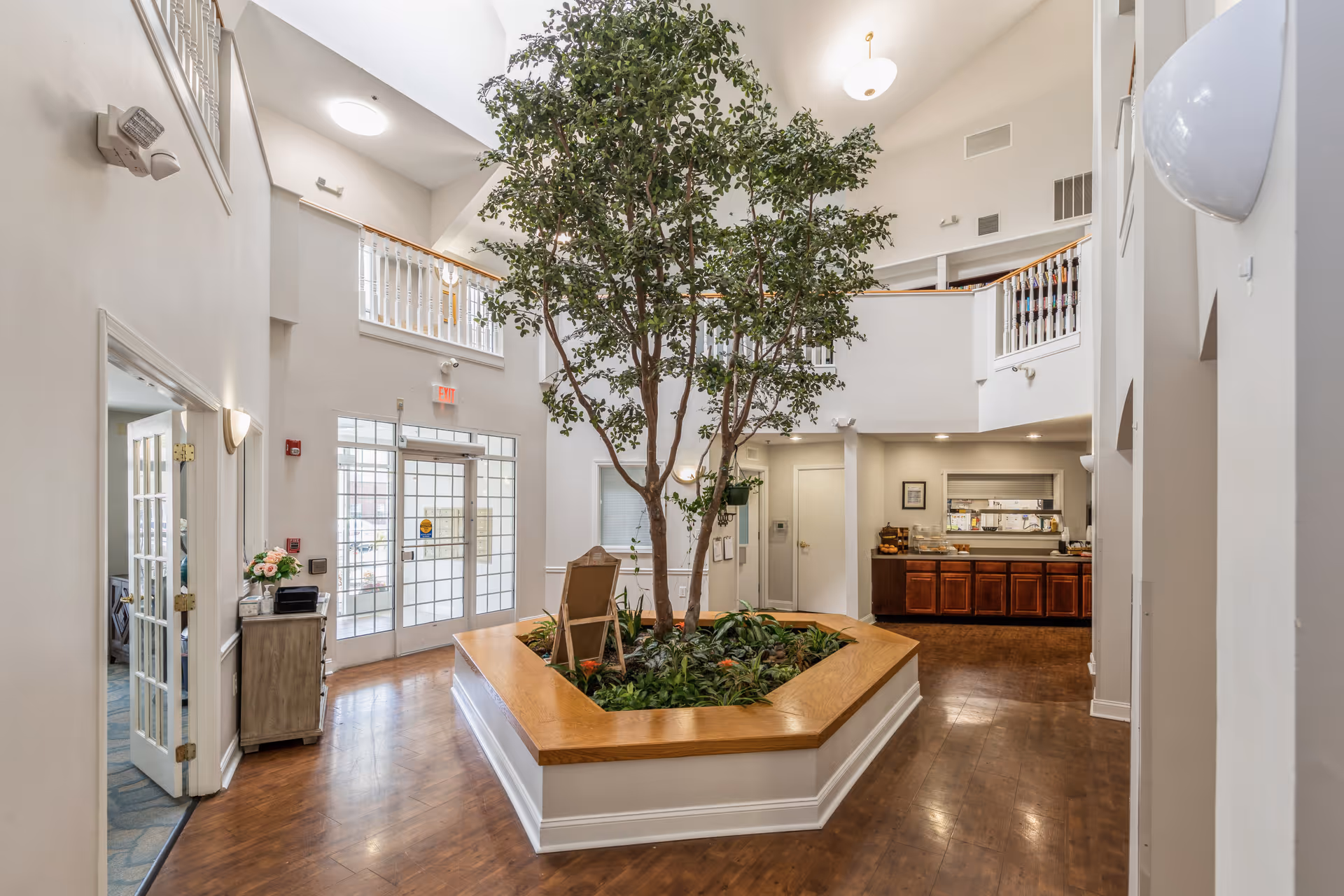 Interior view of a senior living facility lobby with a large indoor tree planted in a wooden planter in the center. The space features high ceilings, white walls, wooden flooring, and a balcony railing on the upper level. There is a glass double door entrance, a small cabinet with flowers on the left, and a counter area with cabinets and a serving window on the right.