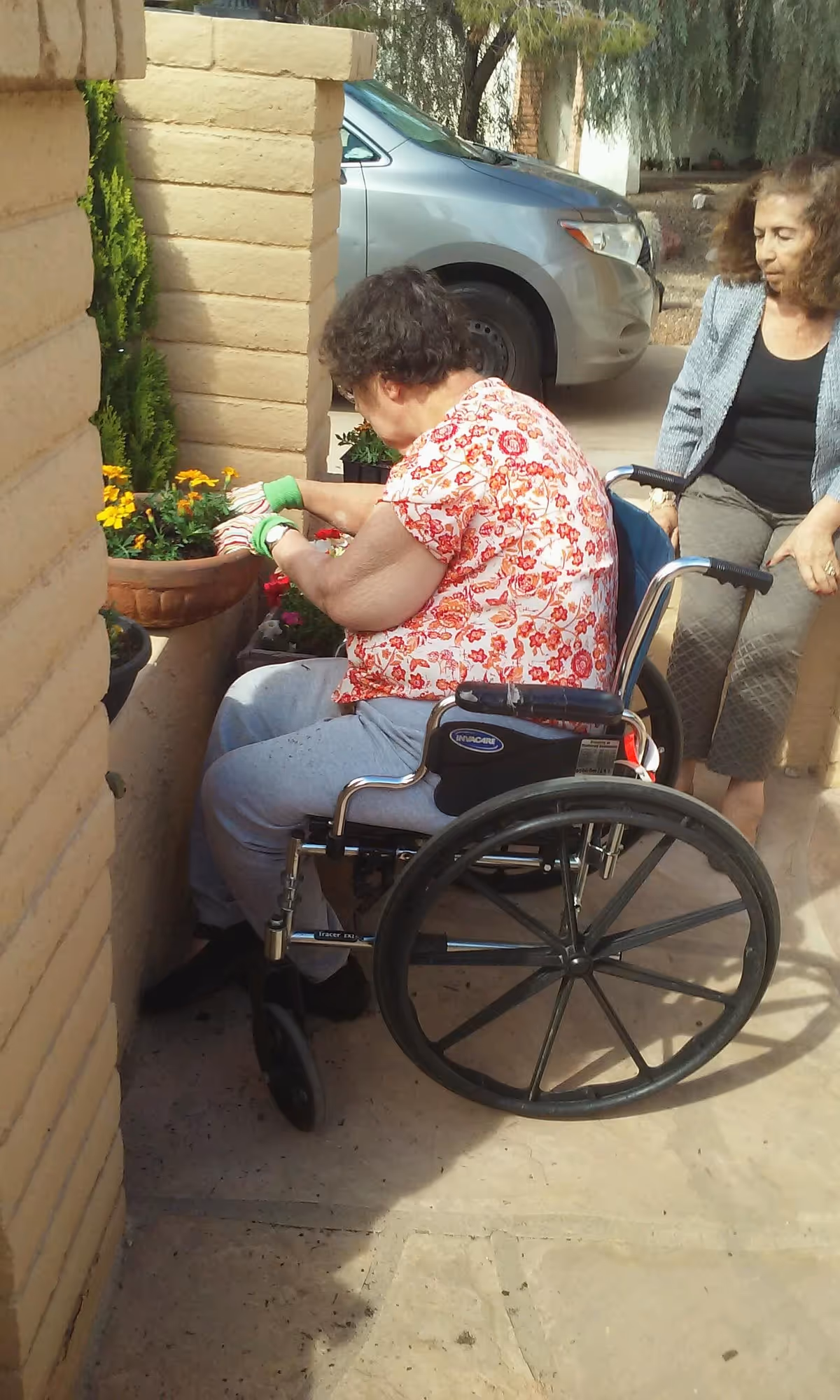 An elderly woman in a wheelchair wearing gardening gloves is tending to flowers in a planter outside, while another woman stands nearby watching. They are in an outdoor area with a car parked in the background.