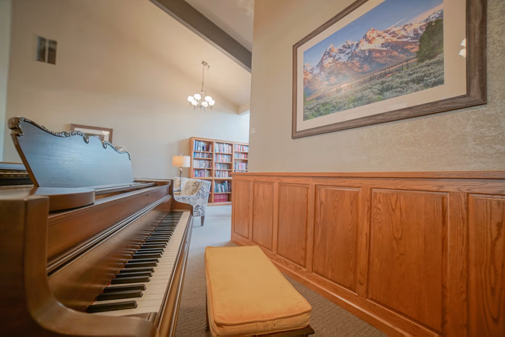 Interior view of a cozy room featuring a wooden piano with a cushioned bench in the foreground. In the background, there is a bookshelf filled with books, a patterned armchair, a floor lamp, and a chandelier hanging from the ceiling. A large framed photograph of a mountain landscape is mounted on the wall above wooden paneling.