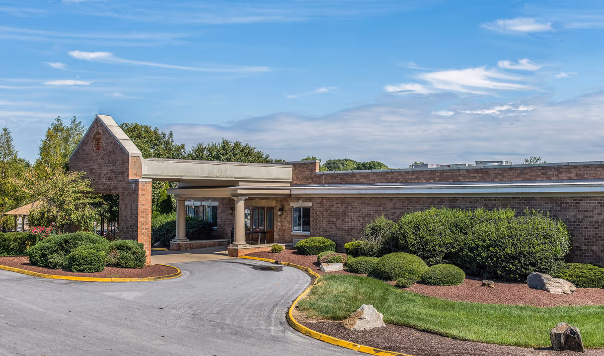 Exterior view of Pike Creek Nursing and Rehabilitation facility showing a brick building with a covered entrance, surrounded by landscaped bushes, grass, and trees under a partly cloudy sky.