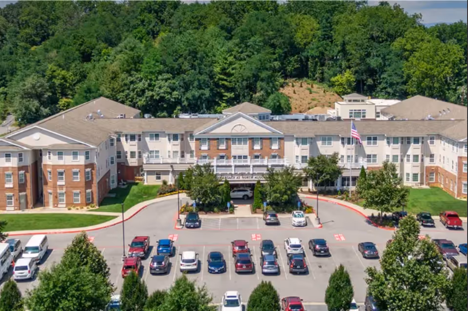 Front exterior of a three-story senior living building with a covered entrance, American flag, and parking lot.