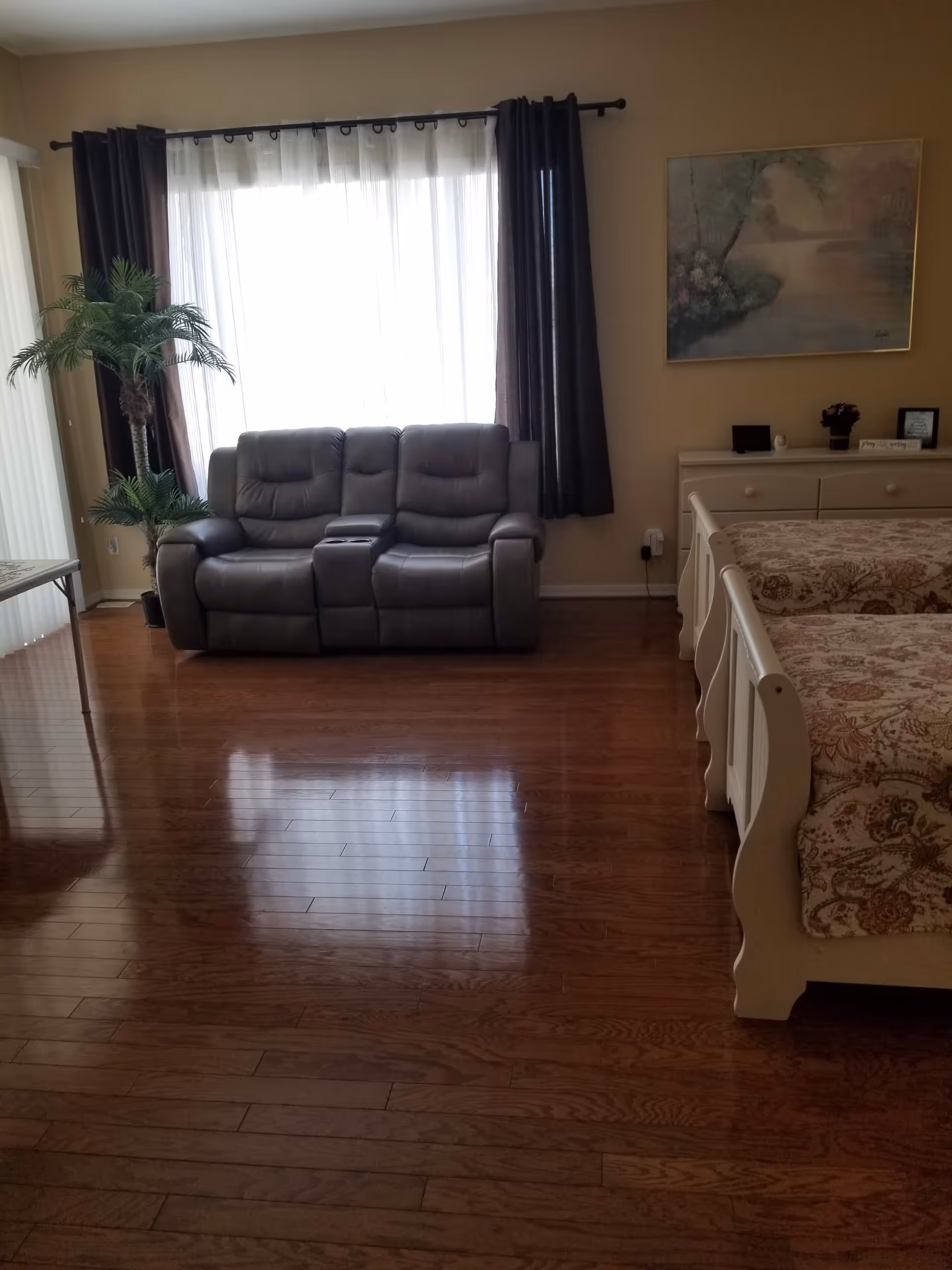 A cozy room with a dark gray leather loveseat positioned in front of a large window with sheer white curtains and dark drapes. To the right, there are two twin beds with patterned bedspreads and a white dresser with decorative items and a framed painting above it. A potted palm plant is placed near the loveseat, and the floor is polished wood.