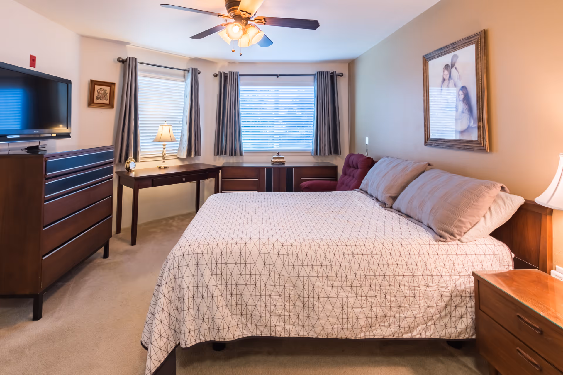 Neatly furnished bedroom with a bed, bedside tables, dresser, TV, desk, and windows with curtains under a ceiling fan.