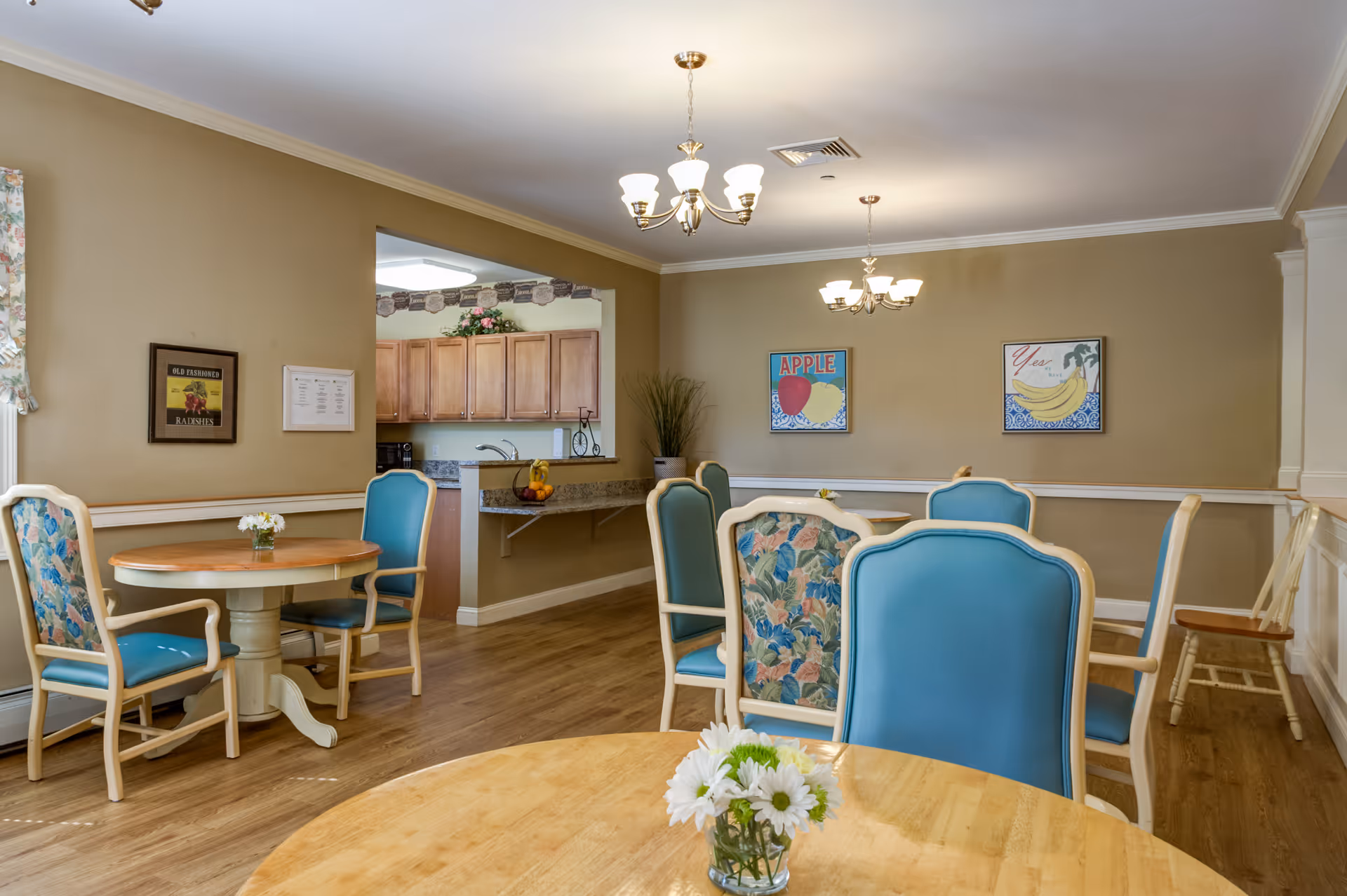 A dining room in a senior living facility with round wooden tables and chairs featuring blue and floral upholstery. The room has beige walls with two framed fruit-themed artworks, a small vase with white flowers on the table in the foreground, and a view into a kitchen area with wooden cabinets. Two chandeliers hang from the ceiling providing warm lighting.