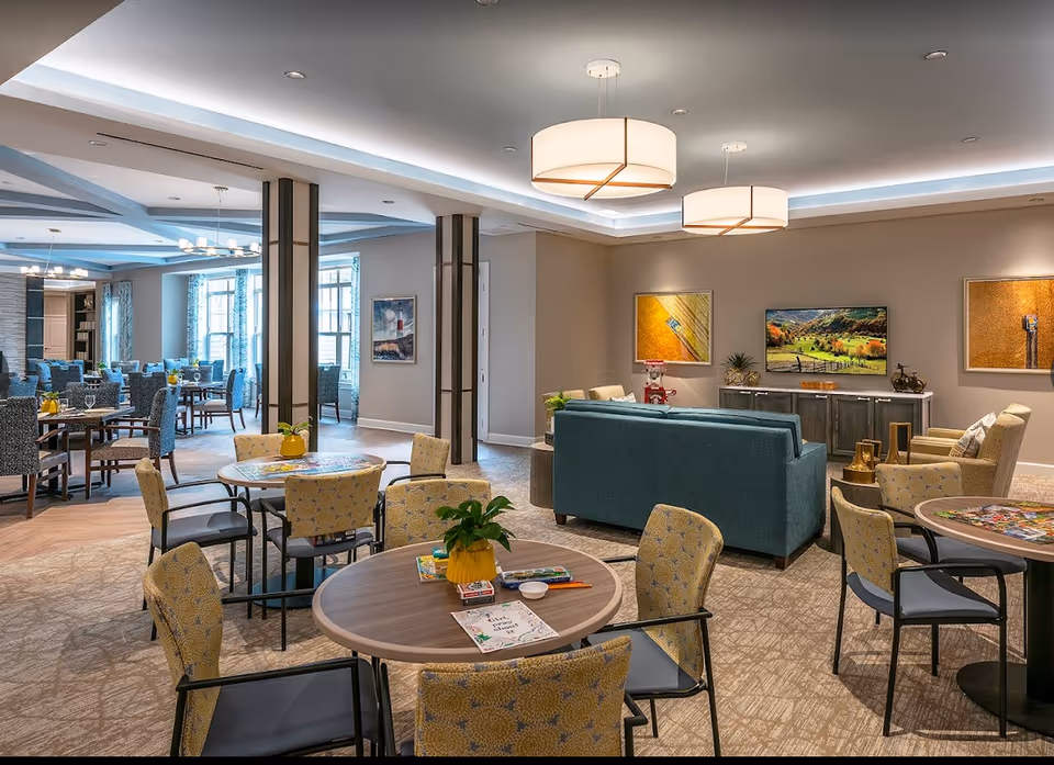 A spacious and well-lit common area in a senior living facility featuring multiple round tables with chairs, a seating area with a teal sofa and beige armchairs, a TV mounted on a cabinet, and colorful artwork on the walls. The room has modern ceiling lights and large windows allowing natural light.