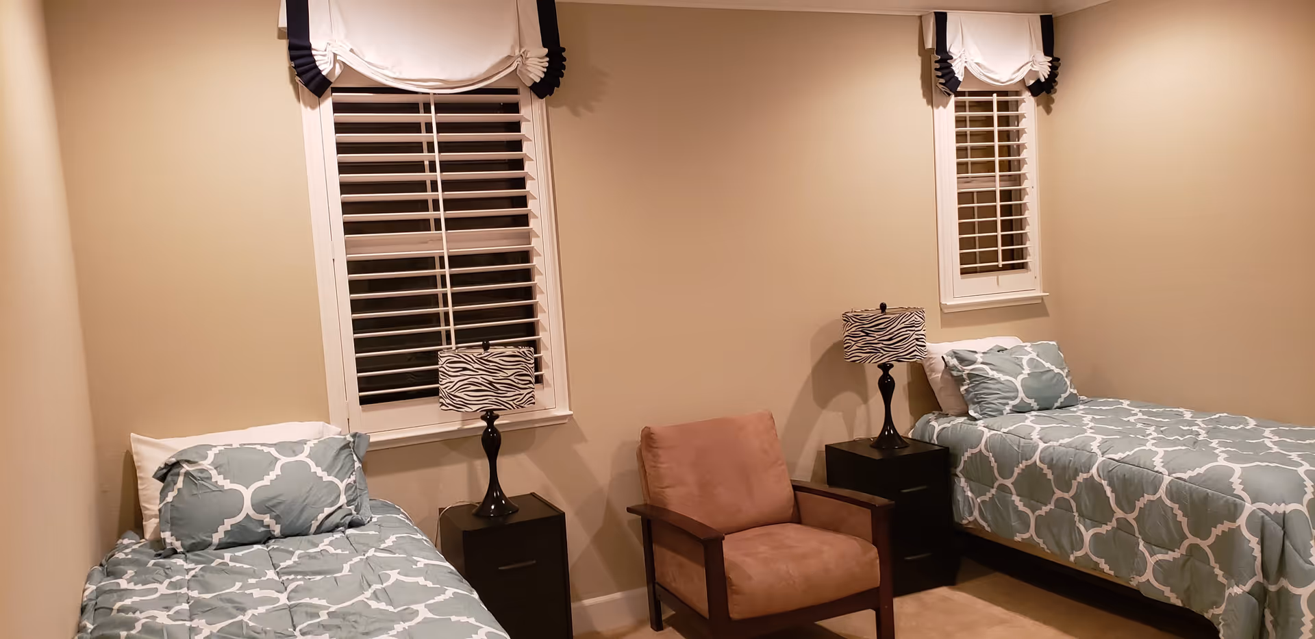 A bedroom with two twin beds, each with a blue and white patterned comforter and pillow. Between the beds are two black nightstands, each with a zebra print lamp. There is a brown cushioned armchair positioned between the beds. The room has beige walls and two windows with white shutters and white curtains with black trim.