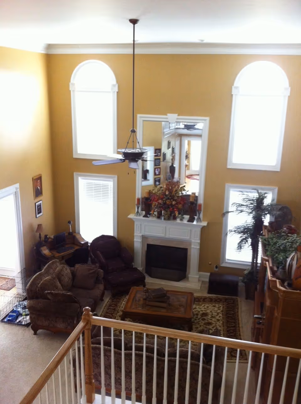 View from an upper level overlooking a living room with beige walls and large arched windows. The room features a ceiling fan, a white fireplace with a large mirror above it, a floral arrangement on the mantel, a brown sofa, a recliner chair, a wooden coffee table on a patterned rug, a small desk with a computer, and various plants and decorations.