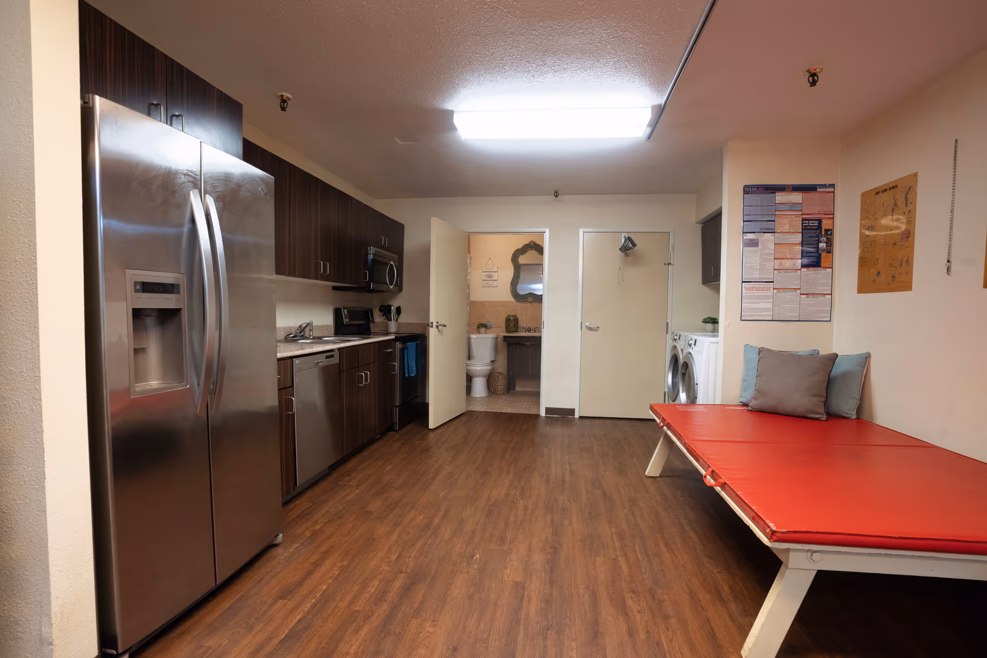 Interior view of a room featuring a modern kitchen with stainless steel appliances including a refrigerator, dishwasher, microwave, and stove. The room has wooden flooring and dark wood cabinets. To the right, there is a red cushioned bench with pillows, a washing machine and dryer, and two doors, one of which leads to a bathroom with a visible toilet and sink. Various posters and notices are on the walls.