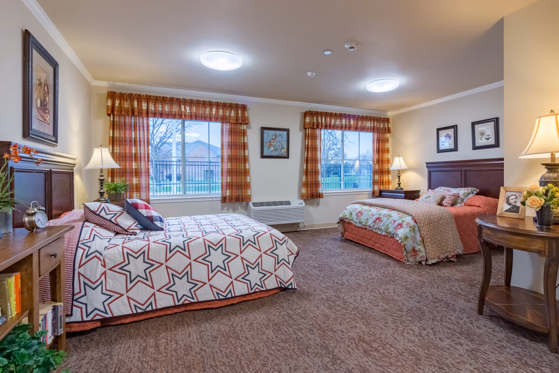 A cozy bedroom in a memory care community featuring two beds with colorful bedding, wooden headboards, and bedside tables with lamps. The room has two large windows with plaid curtains, framed artwork on the walls, and a carpeted floor. There is a small round table with a lamp, flowers, and a framed photo.