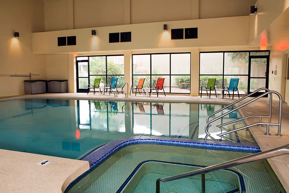 Indoor swimming pool area with a smaller hot tub in the foreground. The pool area has large windows along one wall with colorful chairs lined up outside. The room has beige walls and ceiling with wall-mounted lights and metal handrails for pool access.