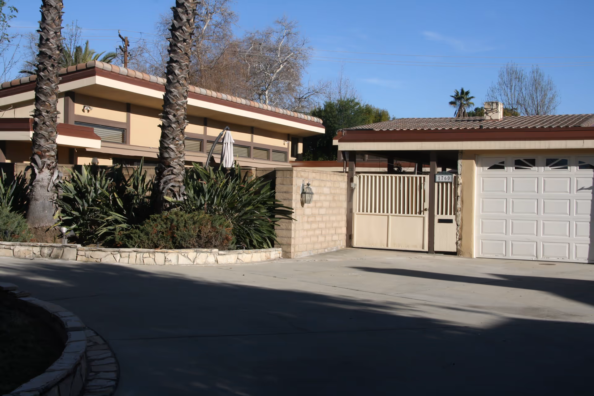 Exterior view of a single-story building with a tiled roof, a white garage door, a gated entrance, and landscaping including palm trees and shrubs under a clear blue sky.