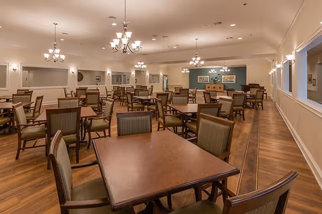 Spacious dining room with multiple wooden tables and cushioned chairs arranged neatly. The room features warm lighting from chandeliers and wall sconces, wood flooring, and light-colored walls with decorative trim. There are windows on one side and framed artwork on the far wall.