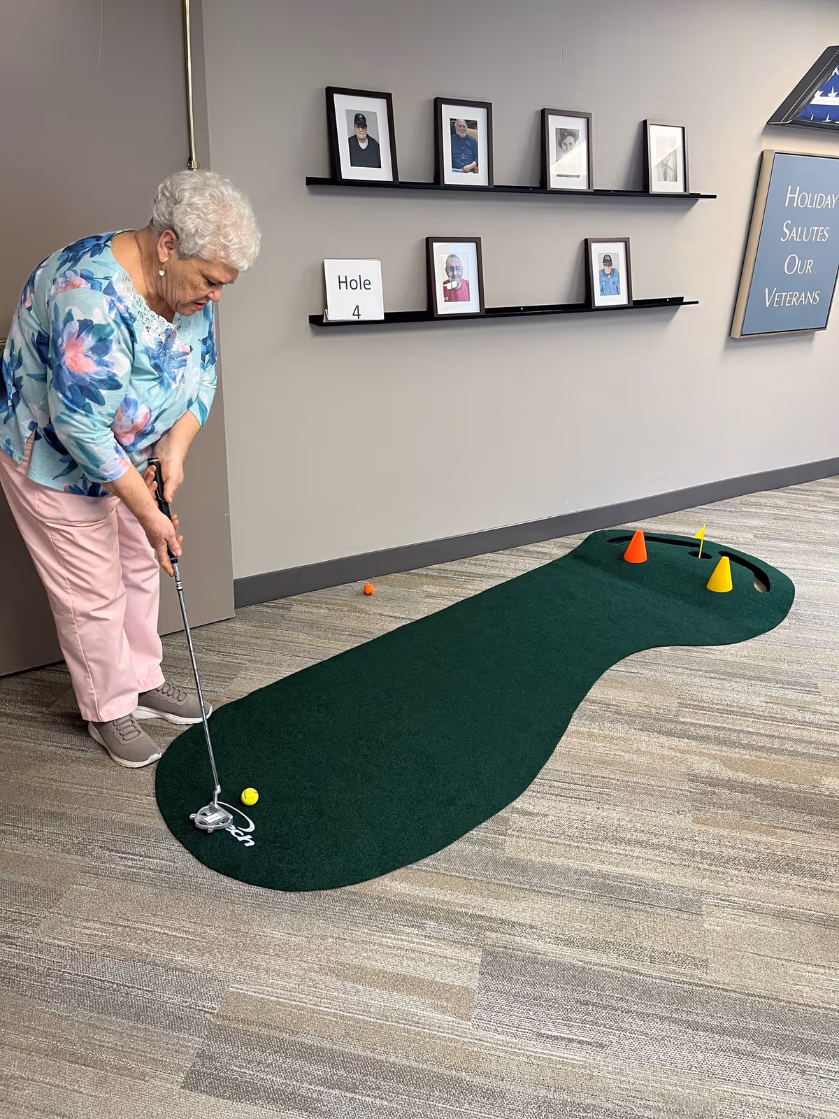 An elderly woman playing indoor mini golf on a green putting mat labeled Hole 4, inside a room with a beige carpet and gray walls. The wall behind her has framed photos and a sign that reads 'Holiday Salutes Our Veterans.'