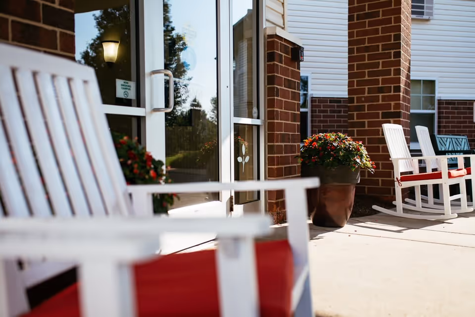 Outdoor seating area at Woodland Village featuring white rocking chairs with red cushions on a concrete patio next to a brick building entrance with glass doors and potted plants with red flowers.