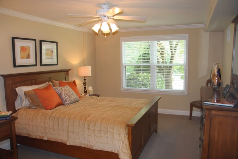 A bedroom with a wooden bed frame and a beige quilted bedspread, decorated with several pillows in orange, beige, and patterned colors. There is a wooden nightstand with a lamp and tissues, two framed pictures on the wall above the bed, a ceiling fan with lights, a window with white blinds showing greenery outside, and a wooden dresser with a chair and decorative items.