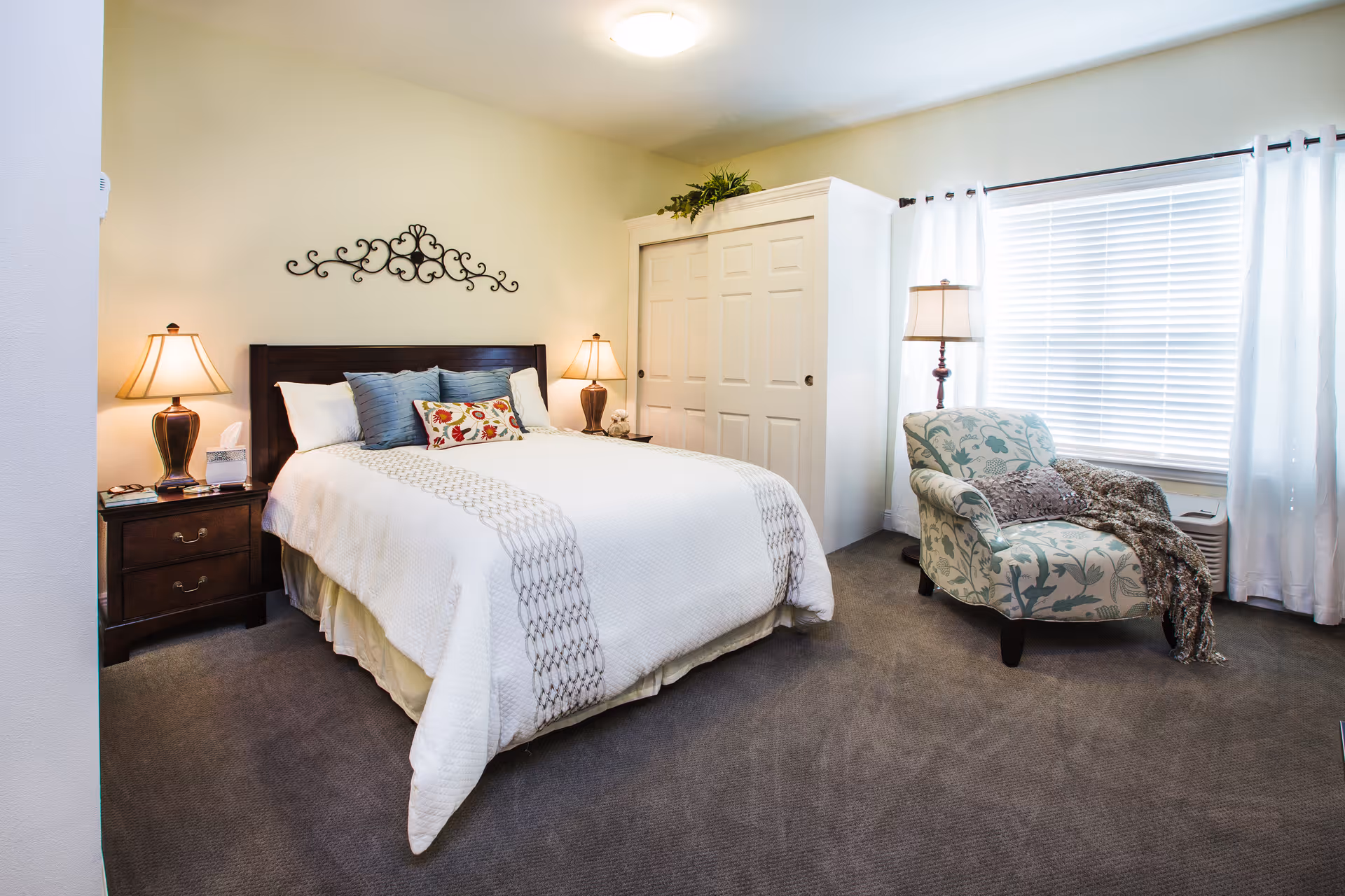 A cozy bedroom with a neatly made bed featuring white bedding and decorative pillows. There are two bedside tables with matching lamps on either side of the bed. A decorative metal wall art piece hangs above the headboard. To the right, there is a patterned armchair with a throw blanket, a floor lamp, and a window with white curtains and blinds. A white closet with sliding doors is visible in the corner.
