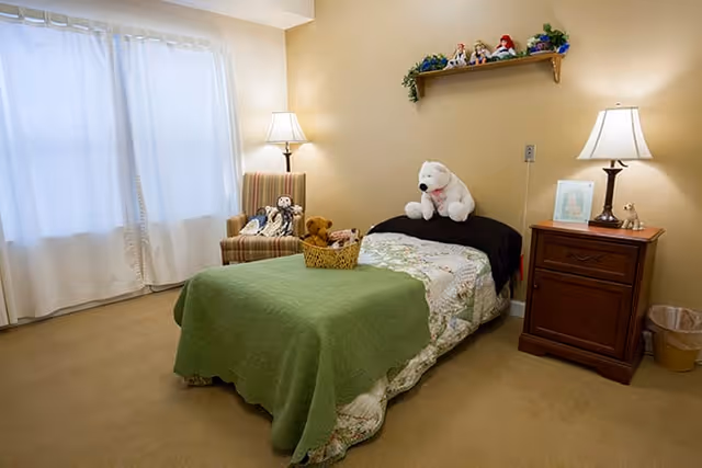 A cozy bedroom with a single bed covered with a green blanket and floral bedspread. There are stuffed animals on the bed and a basket at the foot of the bed. A wooden nightstand with a lamp and framed picture is beside the bed. A striped armchair with more stuffed animals sits near a window with white curtains. A wooden shelf with decorative items is mounted on the beige wall above the bed.