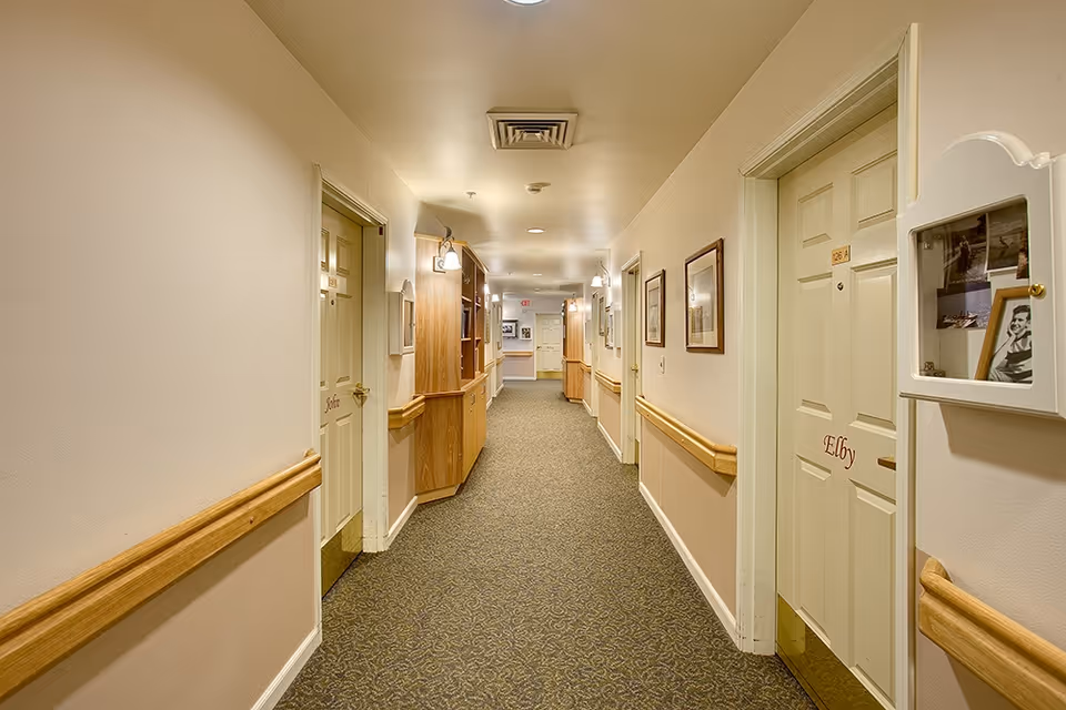 A well-lit hallway in a senior living facility with beige walls, carpeted floor, wooden handrails on both sides, and several closed doors labeled with names. There are framed pictures on the walls and built-in wooden shelves along the left side.