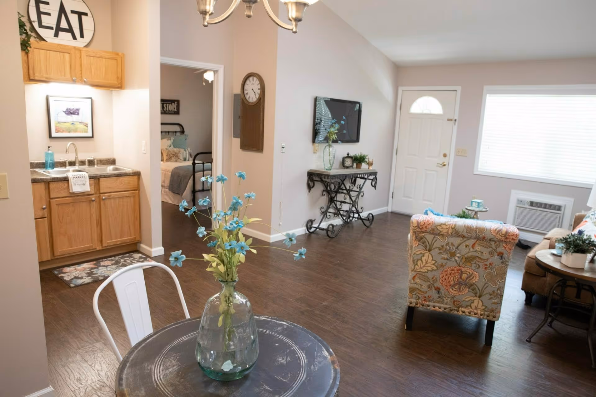 Interior view of a senior living facility showing a small dining table with a glass vase of blue flowers in the foreground, a kitchenette with wooden cabinets and a sink on the left, a bedroom visible through an open doorway, and a living area with a floral patterned armchair, a wall-mounted TV, a decorative table, and a white front door with a window and a large window with blinds.