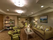 A cozy living room with a beige sofa adorned with patterned cushions, two green armchairs, a glass coffee table with decorative items, a floor lamp, a painting on the wall, and a shelving unit with various decor pieces. The room has warm lighting and wooden flooring.