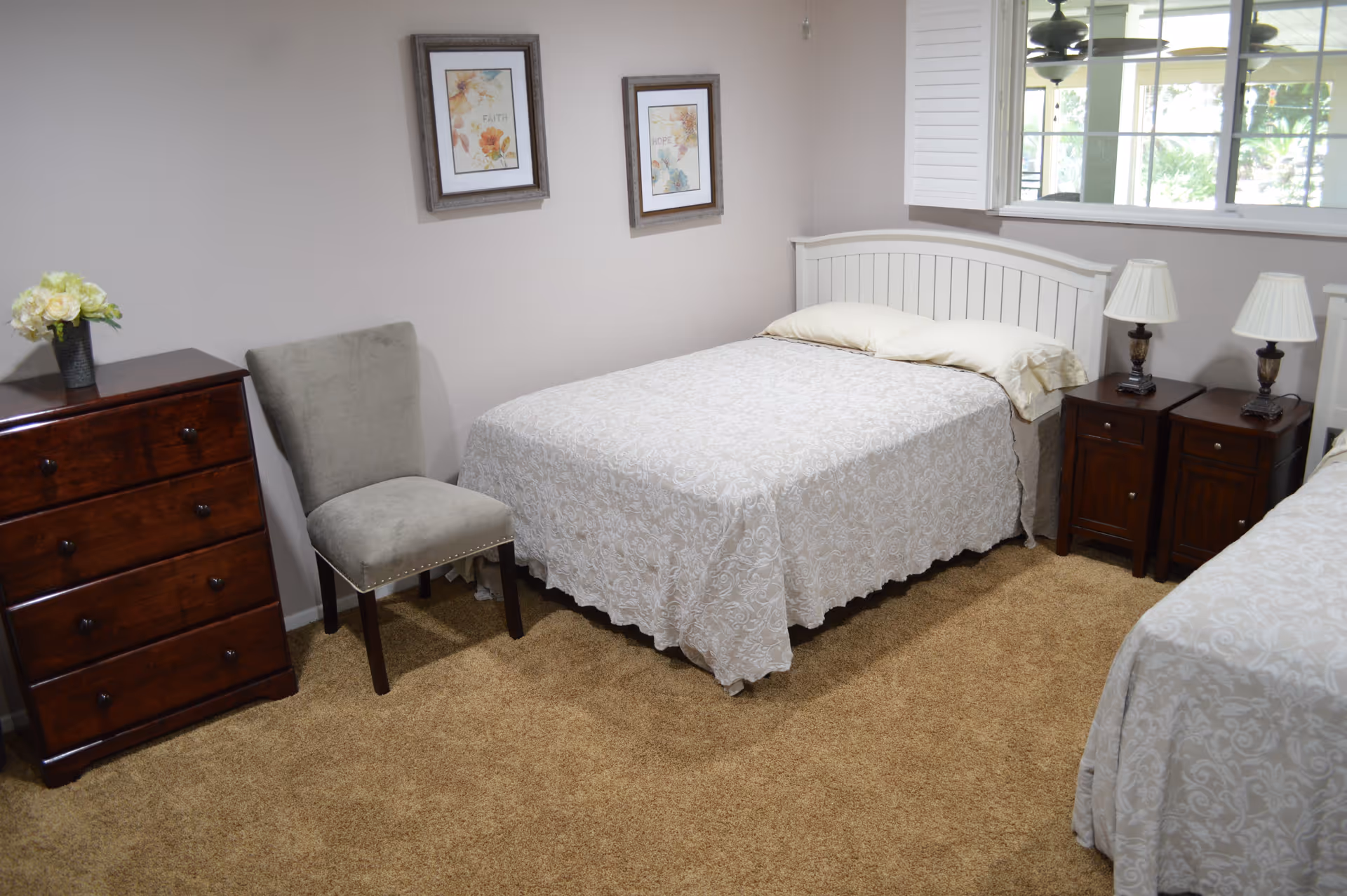A tidy bedroom with two beds, nightstands and lamps, a dresser, a chair, framed artwork, and a window.