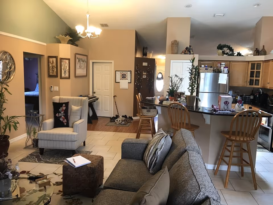 Open-plan living room and kitchen with a sofa and armchair in the foreground and a breakfast bar with wooden stools by the kitchen.