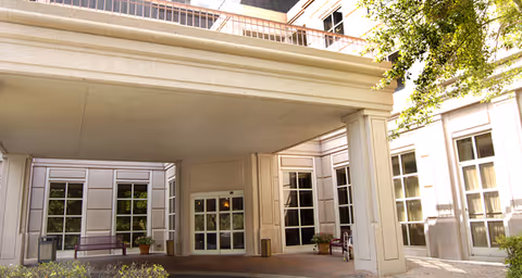 Entrance area of a healthcare facility with a covered drop-off zone supported by large columns. The building has multiple windows and a balcony above the entrance. There are benches and potted plants near the entrance, and trees providing shade on the right side.
