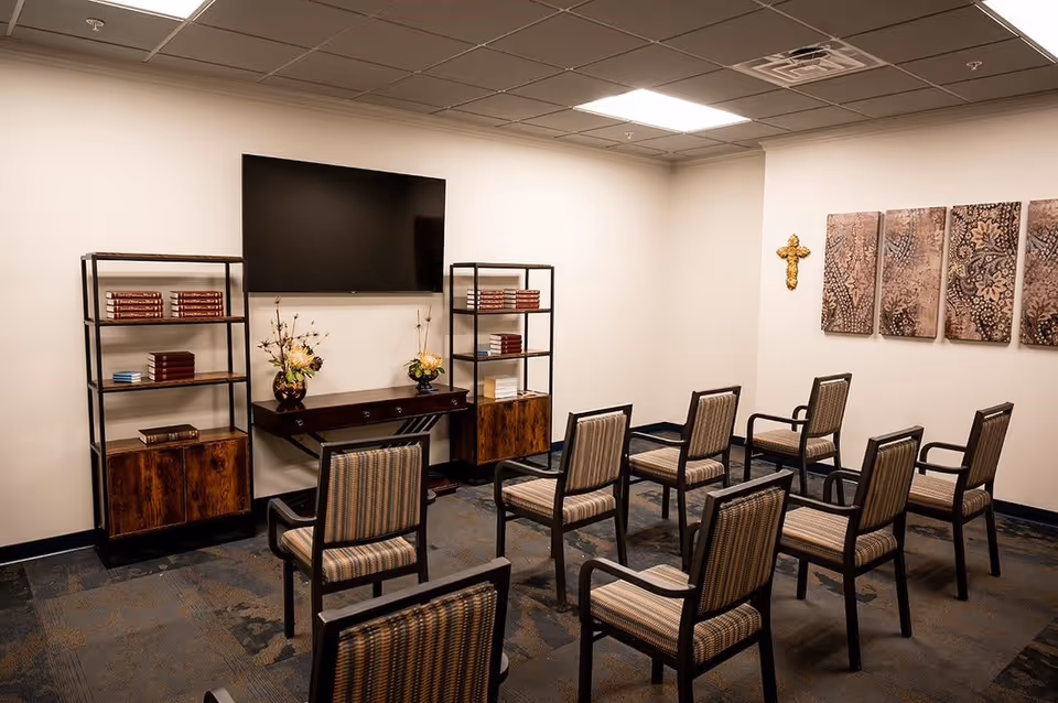 A small meeting or chapel room with nine chairs arranged in rows facing a wall-mounted flat screen TV. Two wooden shelving units with books and flower arrangements flank a small wooden table beneath the TV. On the right wall, there is a decorative cross and four rectangular art pieces with floral patterns. The room has a carpeted floor and a drop ceiling with fluorescent lighting.