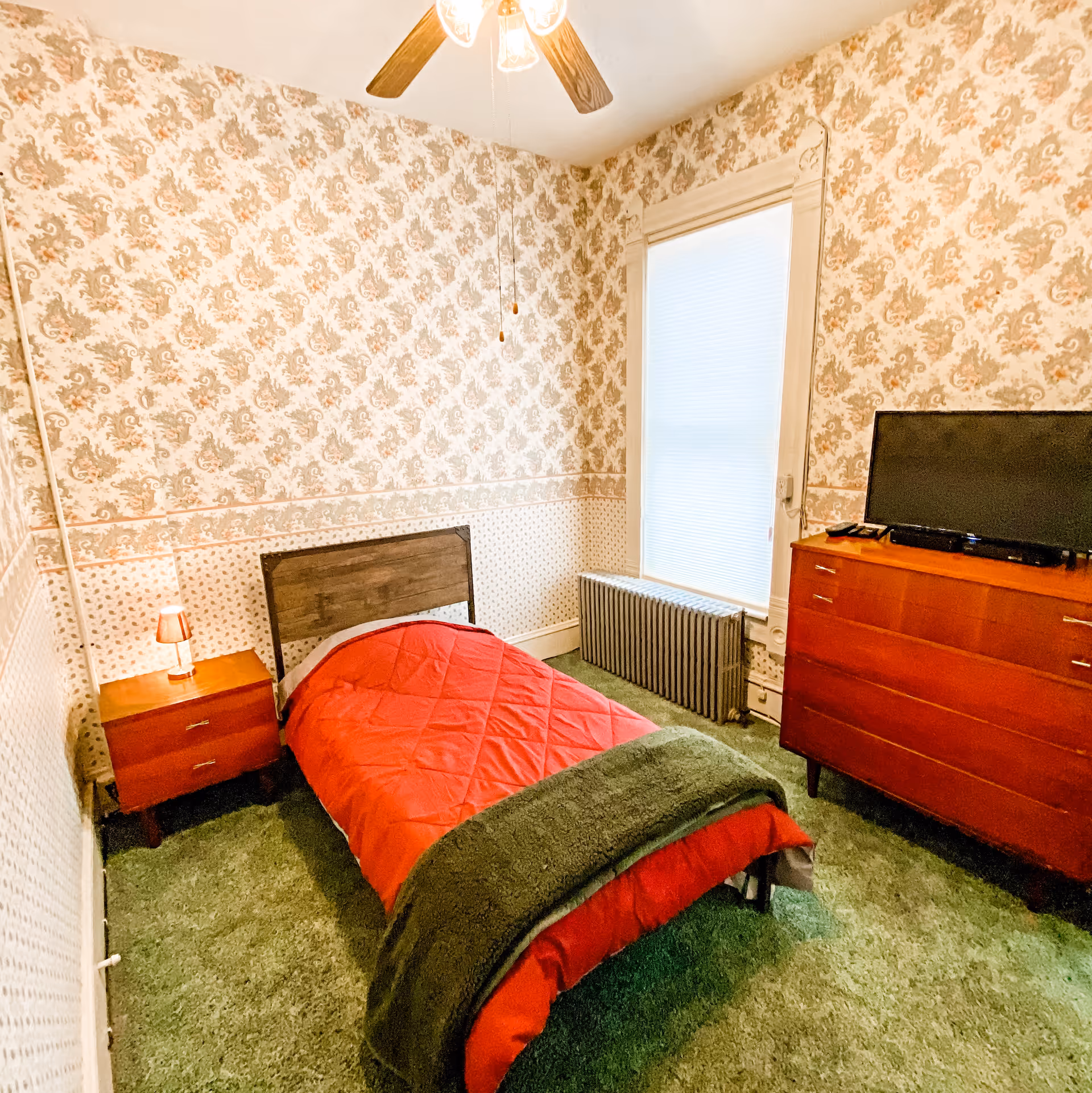 A small bedroom with vintage floral wallpaper and green carpet. The room features a single bed with a red comforter and a green blanket at the foot. There is a wooden nightstand with a small lamp on the left side of the bed and a wooden dresser with a flat-screen TV on top on the right side. A window with a white blind and a radiator underneath is on the far wall. A ceiling fan with wooden blades is visible above.