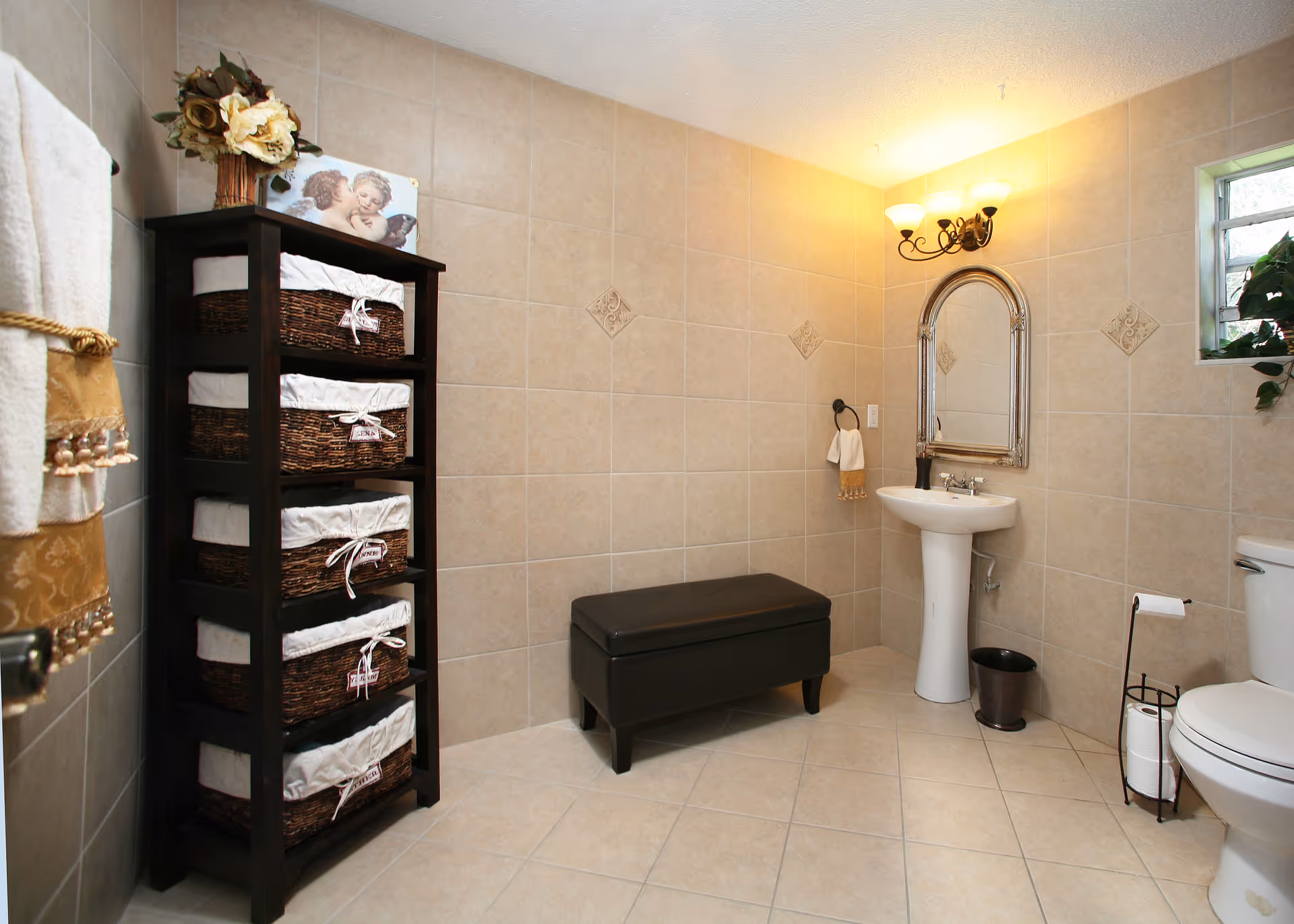 A clean and well-lit bathroom with beige tiled walls and floor. The room features a white pedestal sink with a decorative mirror above it, a black cushioned bench, a toilet with a metal toilet paper holder, and a tall dark wooden shelving unit with wicker baskets. There is a small window with a plant on the right side and a wall-mounted light fixture above the mirror.